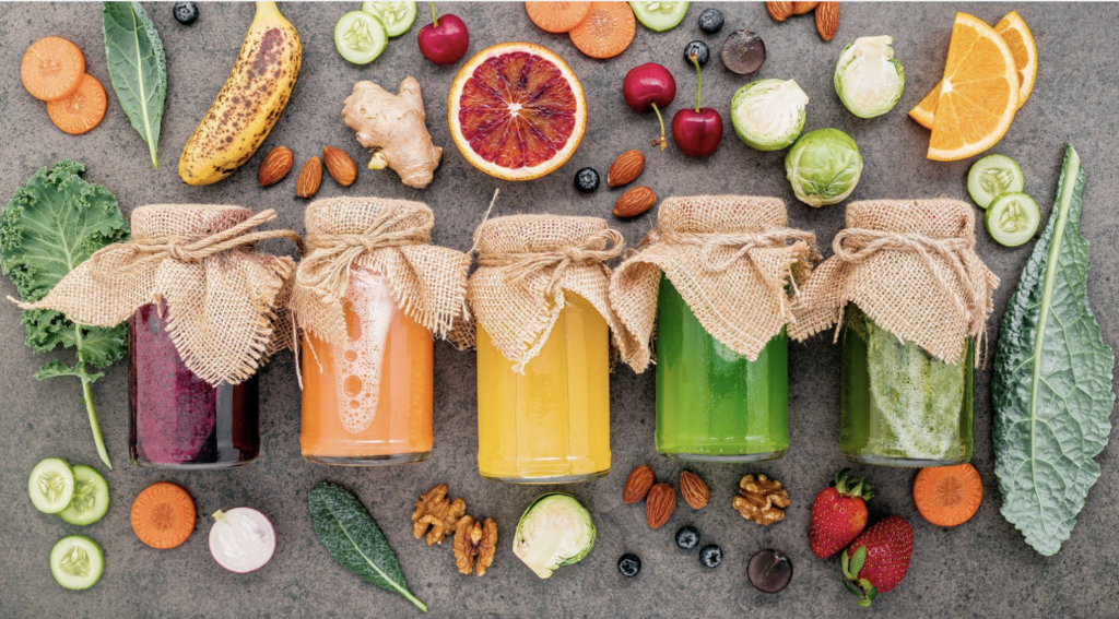 There are five juices in glass jars in the middle of the image. Each a different color. Surrounding the juices are a variety of fruits and vegetables to use for juicing.